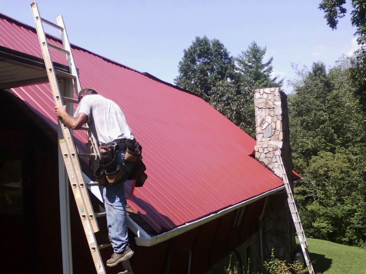Roofer on a ladder beside a red metal roof and stone chimney — Soffit & Fascia Repair by Roofing Brothers in Gresham